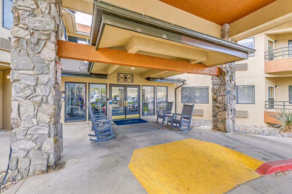 Covered entrance area of a senior living facility with stone pillars and a yellow painted curb. There are three blue rocking chairs placed near the glass double doors leading inside the building. The building exterior is beige with multiple windows and balconies.