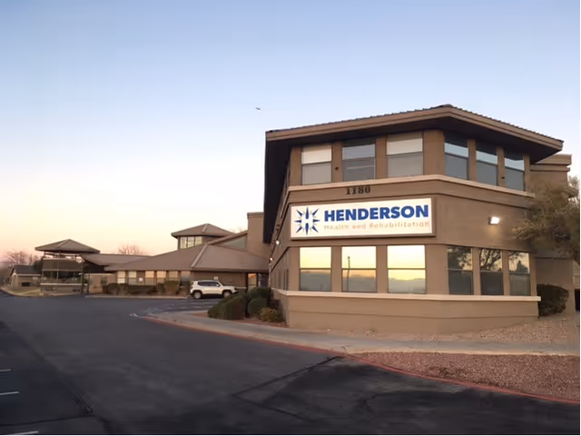 Exterior view of Henderson Health and Rehabilitation building at sunset, showing a two-story structure with large windows and a sign displaying the facility's name. The surrounding area includes a paved driveway and some landscaping with bushes and gravel.