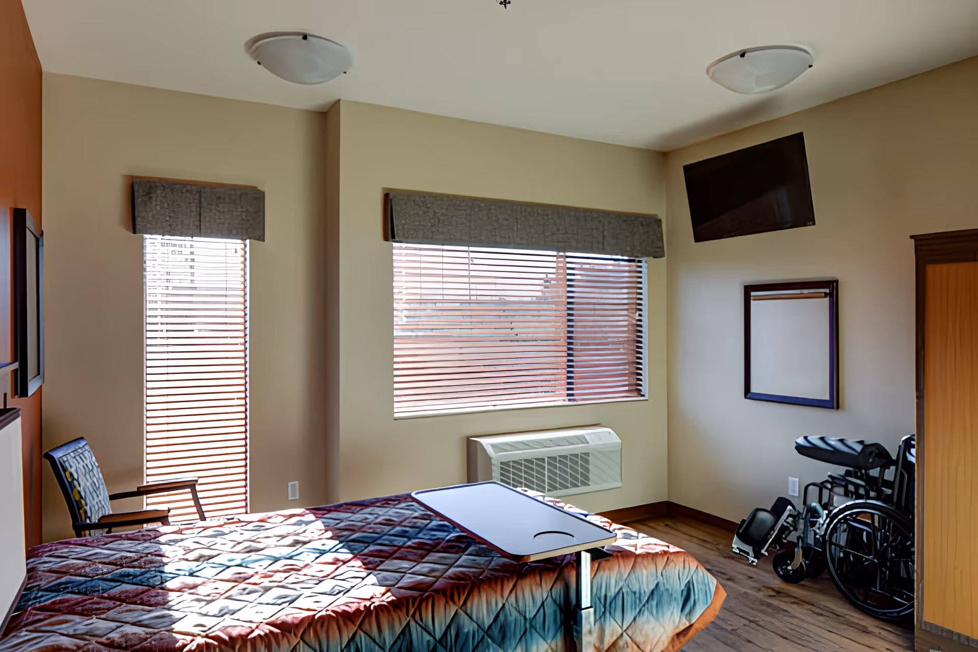 A bedroom in a senior living facility with a bed covered by a colorful quilt, a small overbed table, a chair near a window with blinds, a wall-mounted TV, a wheelchair, and a wooden wardrobe. The room has beige walls and wooden flooring.