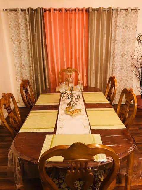 A wooden dining table with six chairs, placemats, and a decorative centerpiece set before patterned curtains.
