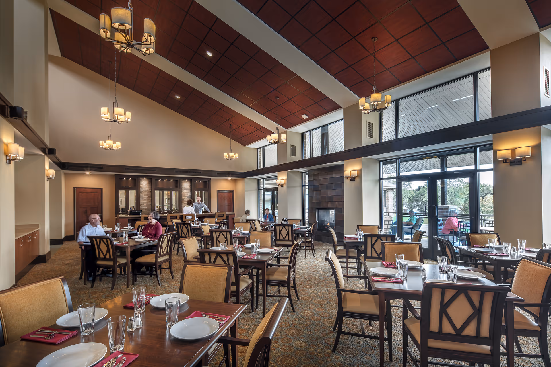 A spacious dining room with high ceilings and large windows allowing natural light. Several tables are set with plates, glasses, and red napkins. A few elderly people are seated and engaged in conversation, while a staff member stands near the back. The room features warm lighting fixtures, a fireplace, and a view of an outdoor patio area.