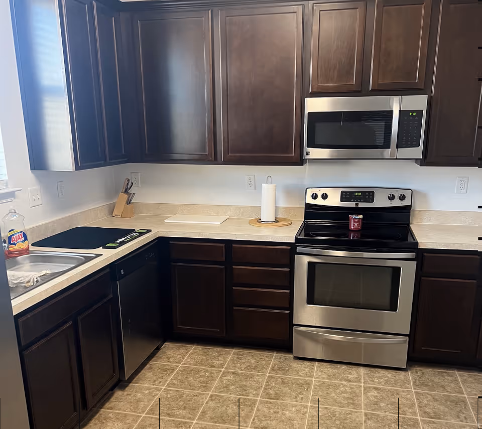 A kitchen with dark wooden cabinets, a stainless steel microwave and oven, a dishwasher, a sink with a bottle of Ajax dish soap, a paper towel holder, a knife block, and a cutting board on the countertop.
