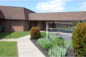 Exterior view of a single-story brick building with a covered walkway supported by columns. There is a paved driveway and landscaped garden beds with bushes and plants along the sidewalk leading to the entrance.