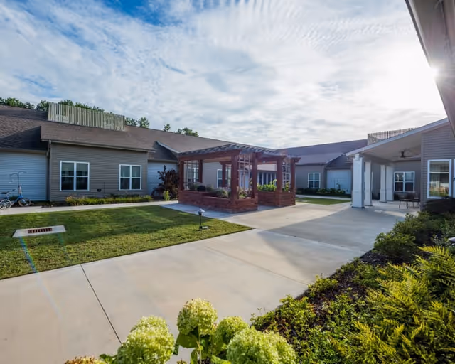 Outdoor courtyard area of Clear Creek Memory Care facility featuring a paved walkway, green lawn, a wooden pergola with brick base, surrounding single-story building with windows, and some landscaping with bushes and flowers under a partly cloudy sky.