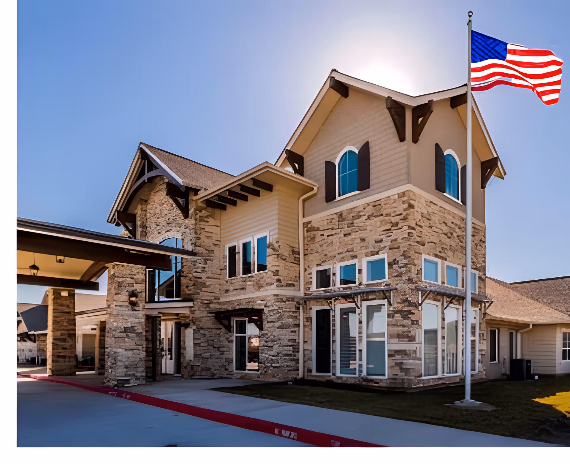 Exterior view of a senior living facility building with stone and beige siding, multiple windows, and a peaked roof. An American flag is flying on a flagpole in front of the building under a clear blue sky.