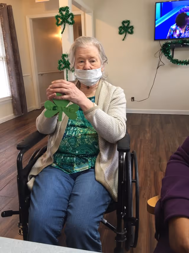 An elderly woman wearing a face mask sits in a wheelchair inside a room decorated with green shamrocks. She is holding a green shamrock decoration. The room has wooden flooring, a TV mounted on the wall, and a window with curtains.