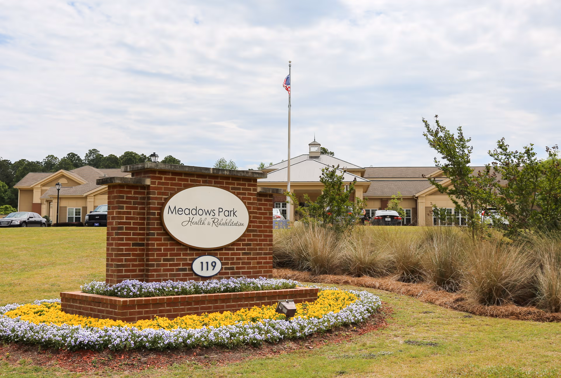 Brick entrance sign for Meadows Park Health & Rehabilitation in front of the facility building with landscaped flowers and a flagpole.