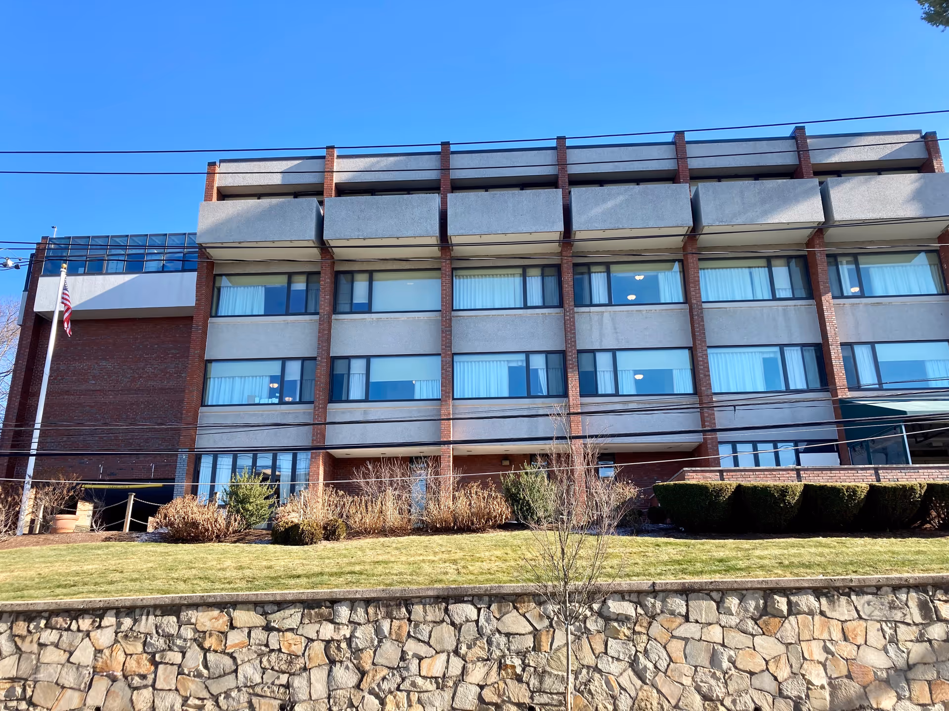Exterior view of a multi-story building with large windows and balconies, a stone retaining wall in front, some bushes and grass, and a flagpole with an American flag on the left side under a clear blue sky.