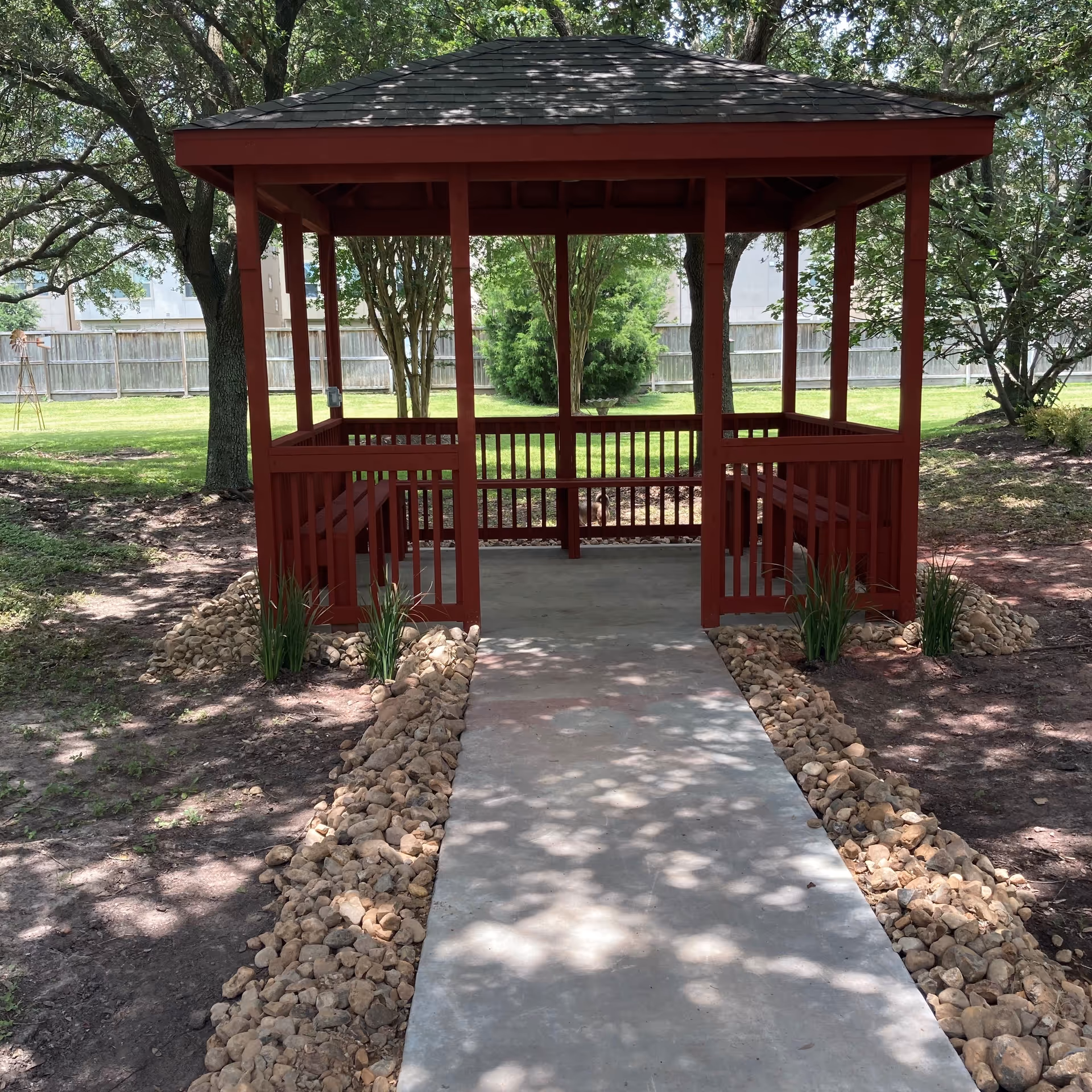 A red wooden gazebo centered at the end of a concrete path in a shaded grassy yard with trees.