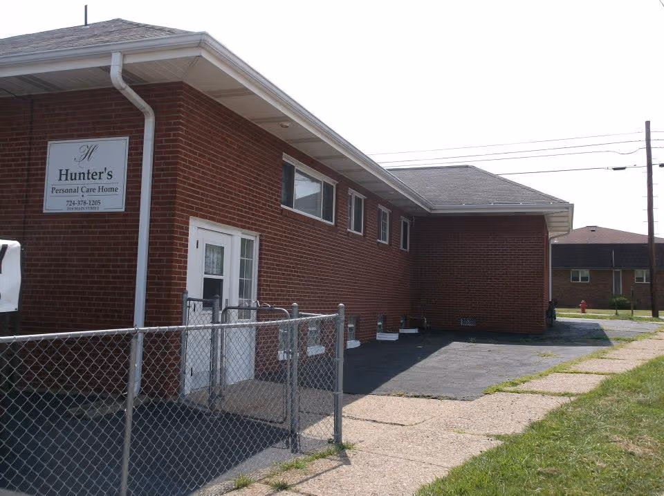 Exterior view of a single-story brick building with a sign that reads 'Hunter's Personal Care Home'. The building has several windows and a white door with a small fenced area in front. There is a paved driveway and a sidewalk with grass alongside it.