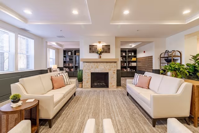 A bright and spacious living room with two cream-colored sofas facing each other across a large beige rug. The room features a stone fireplace in the center with decorative wall art and a plant above it. Large windows on the left side allow natural light to fill the space. Bookshelves and a mail area are visible in the background, along with various plants and a wooden side table with a small plant and books.