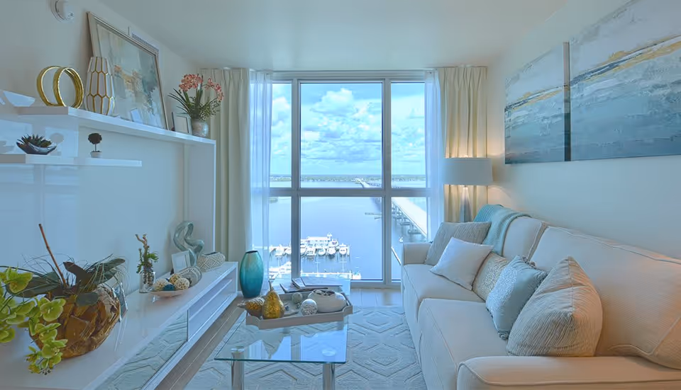 A bright living room with a large window offering a view of a marina and a bridge over water. The room features a beige sofa with multiple cushions, a glass coffee table with decorative items, a white entertainment unit with plants and art pieces, and a floor lamp beside the sofa. Light curtains frame the window.