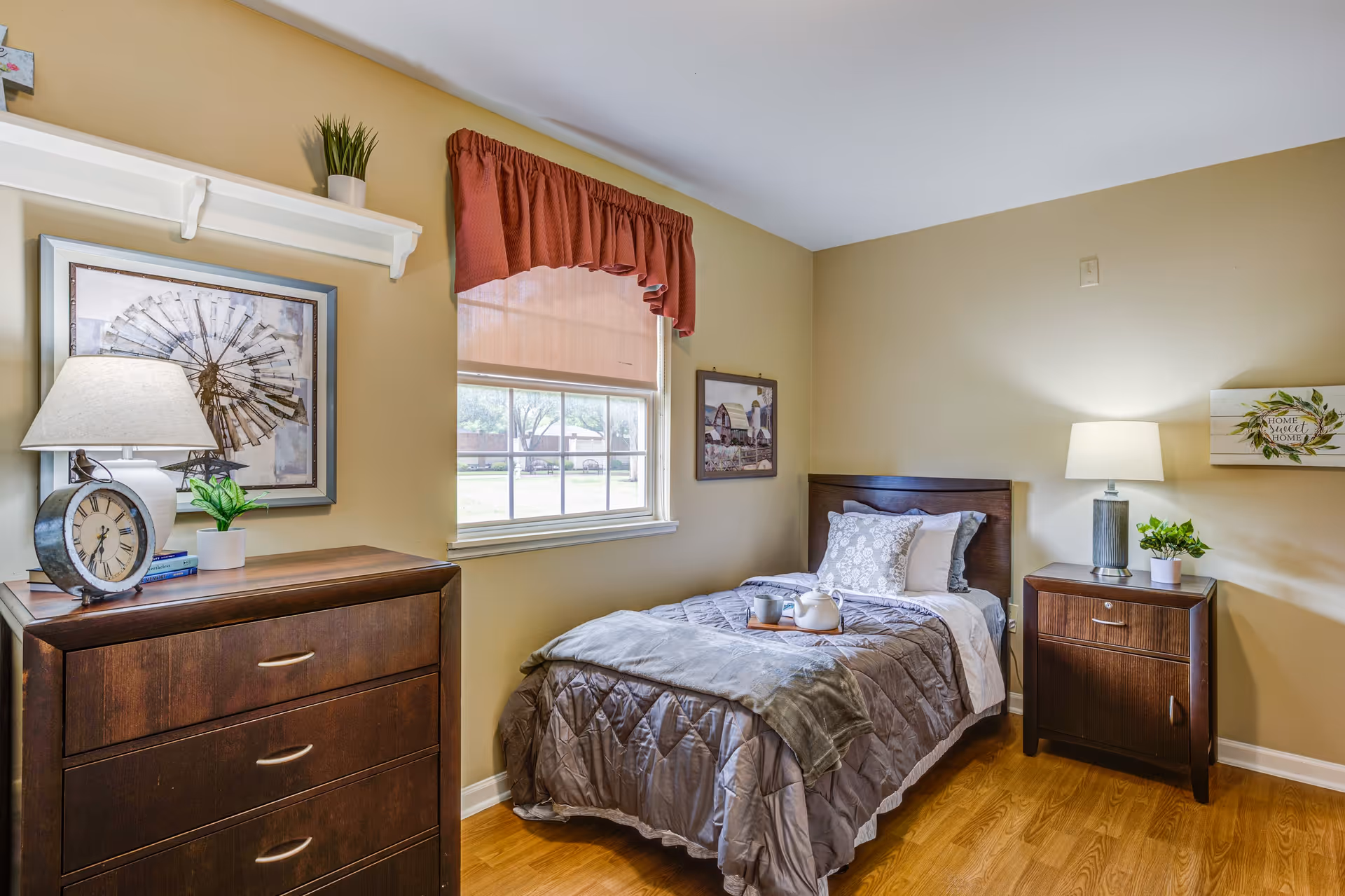 A cozy bedroom with a single bed made with gray bedding and decorative pillows. A wooden nightstand with a lamp and a small plant is beside the bed. Across from the bed is a wooden dresser with a clock, a lamp, and a small plant on top. The room has beige walls, a window with a red valance, and framed artwork on the walls.