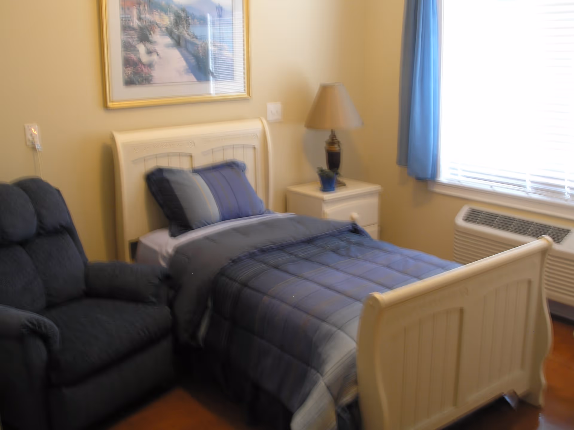 A cozy bedroom with a single bed made with blue and gray bedding, a matching pillow, a white nightstand with a lamp and a small plant, a dark upholstered armchair, a window with blue curtains, and a framed picture on the wall.
