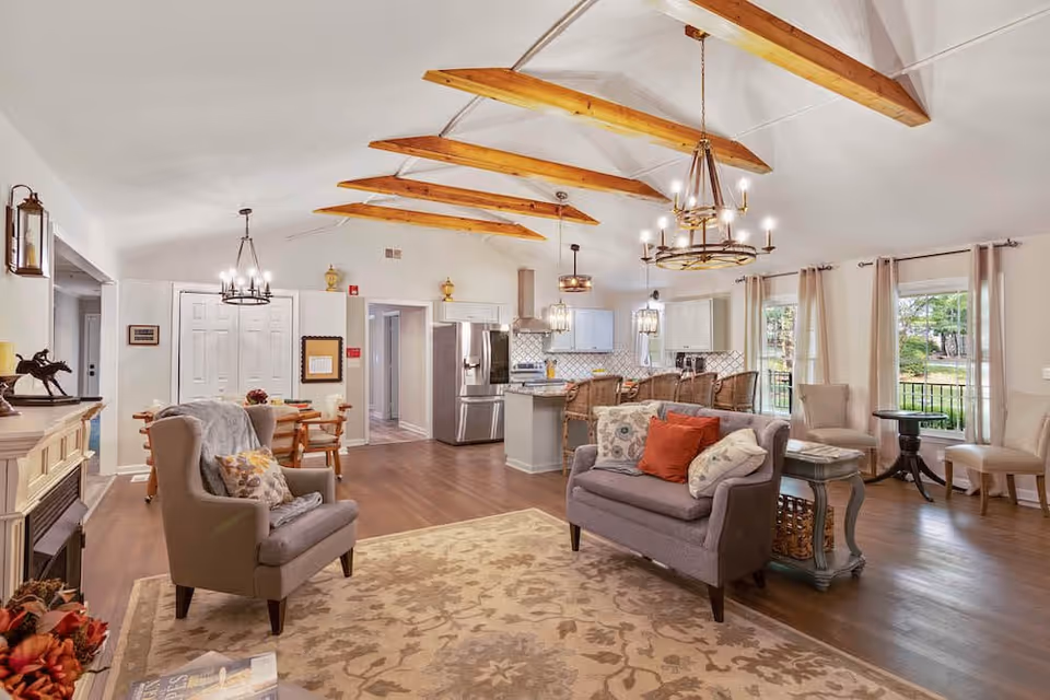 Open, airy living room and kitchen with a vaulted ceiling and exposed wooden beams, upholstered seating, area rug, dining table and a central kitchen island.