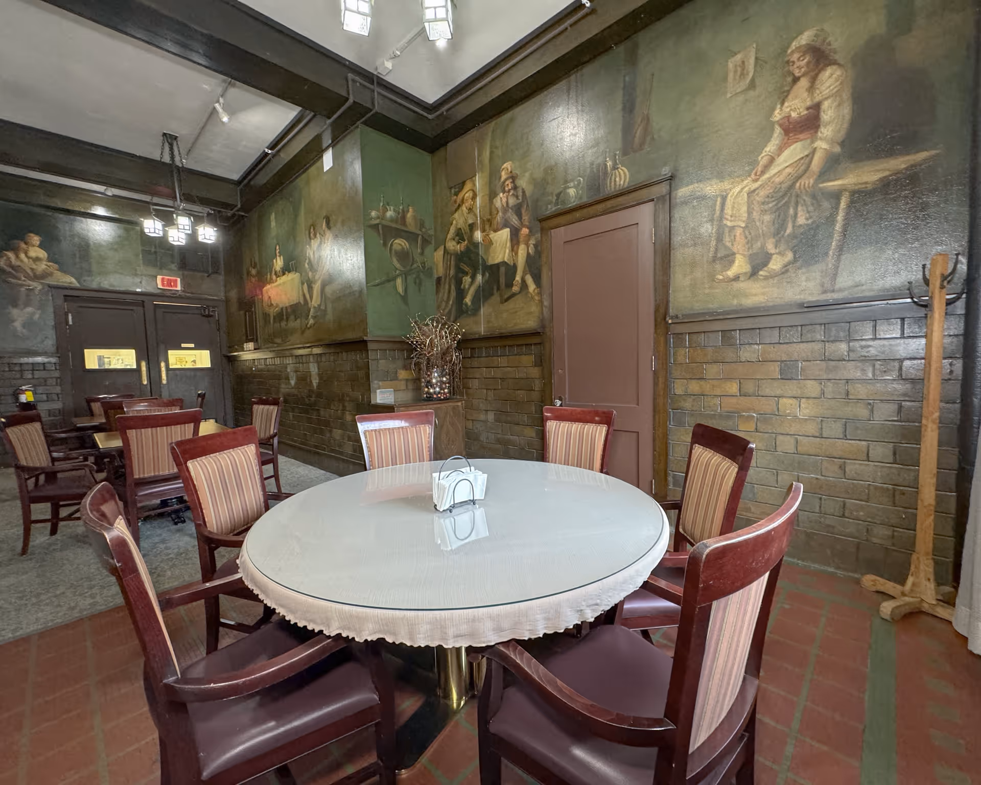 Interior dining room with a round table covered with a white cloth and six wooden chairs with striped upholstery. The walls are decorated with large vintage-style paintings depicting people in historical attire. The room has a tiled lower wall section and a high ceiling with hanging lights. There is a coat rack in the corner and double doors with windows at the back.