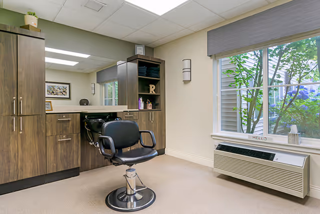 A small hair salon area inside a senior living facility with a black salon chair in front of a counter and sink. There are wooden cabinets and shelves with hair care products, a large window with a view of greenery outside, and a wall-mounted air conditioning unit below the window.