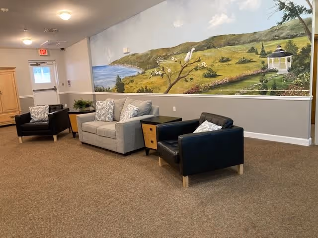 A cozy lounge area with a gray sofa and two black armchairs arranged around a small wooden side table. The wall behind the seating features a large mural depicting a scenic landscape with hills, a lake, trees, flowers, and a white gazebo. The room has beige carpeting and a door with an exit sign above it.
