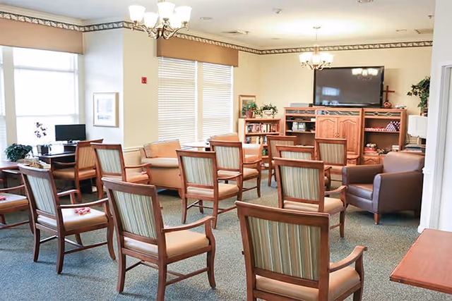 A bright and spacious senior living common area with multiple wooden chairs arranged in rows facing a large flat-screen TV mounted on a wooden entertainment center. The room has beige walls, carpeted floor, large windows with blinds, and two chandeliers providing overhead lighting. There are also a few armchairs and a small desk with a computer in the corner.