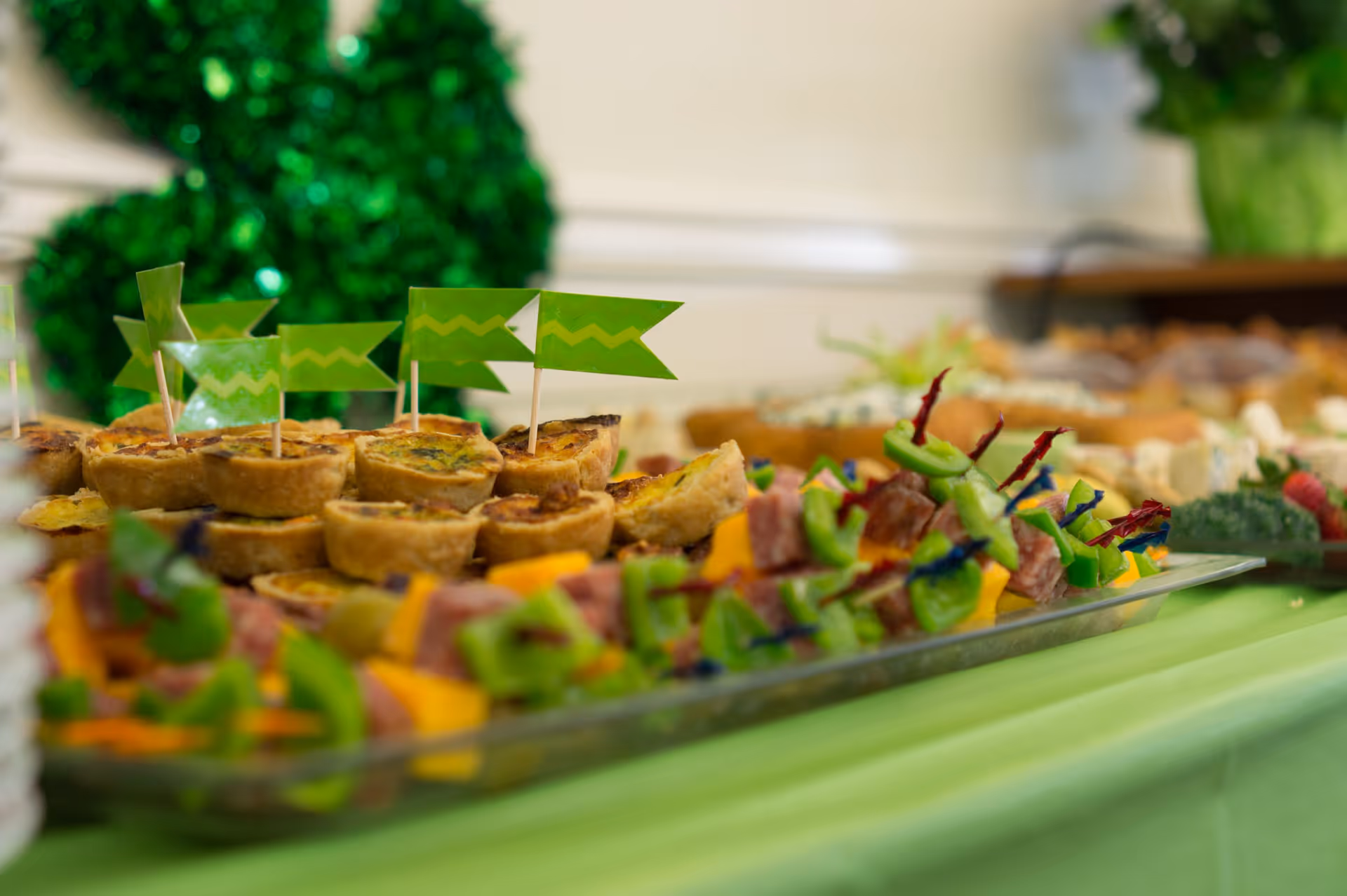 Close-up view of a buffet table with assorted appetizers including mini quiches with green flag toothpicks and skewers with vegetables and meats, set on a green tablecloth with blurred greenery decorations in the background.