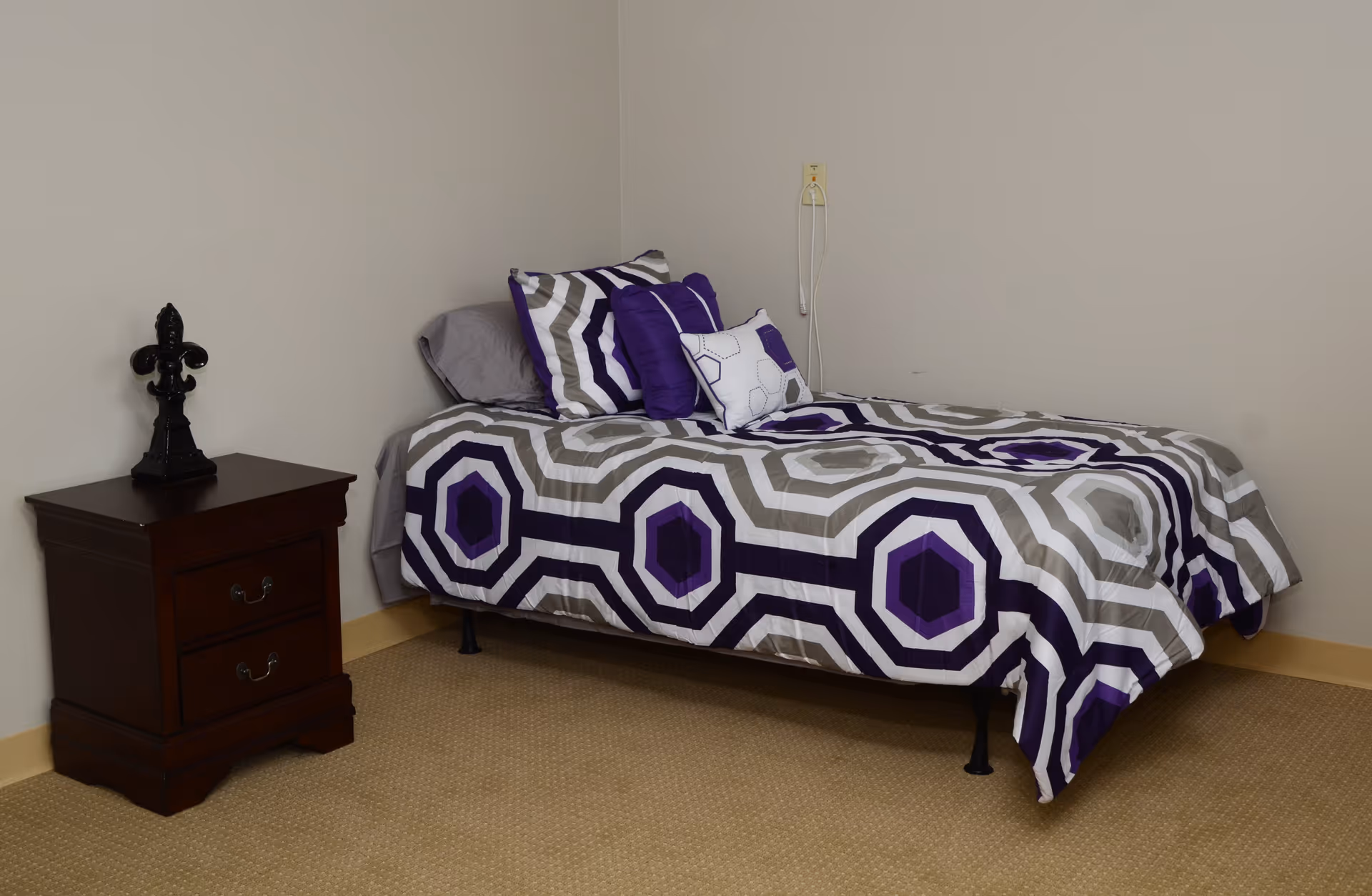 A single bed with a geometric patterned bedspread in shades of purple, gray, and white, accompanied by matching pillows. Next to the bed is a dark wooden nightstand with two drawers and a decorative black fleur-de-lis ornament on top. The room has beige carpet and plain light-colored walls.