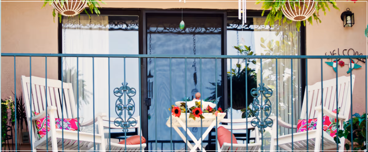 A cozy outdoor patio area with two white rocking chairs featuring colorful cushions, a small white table with a flower arrangement, hanging plants above, and a sliding glass door in the background.