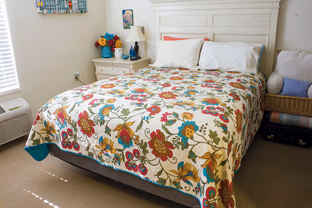A bedroom with a bed covered in a colorful floral quilt. The bed has white pillows and a white headboard. Next to the bed is a nightstand with a lamp, flowers, and decorative items. There is a window with blinds on the left side and a wicker chair with cushions on the right side.