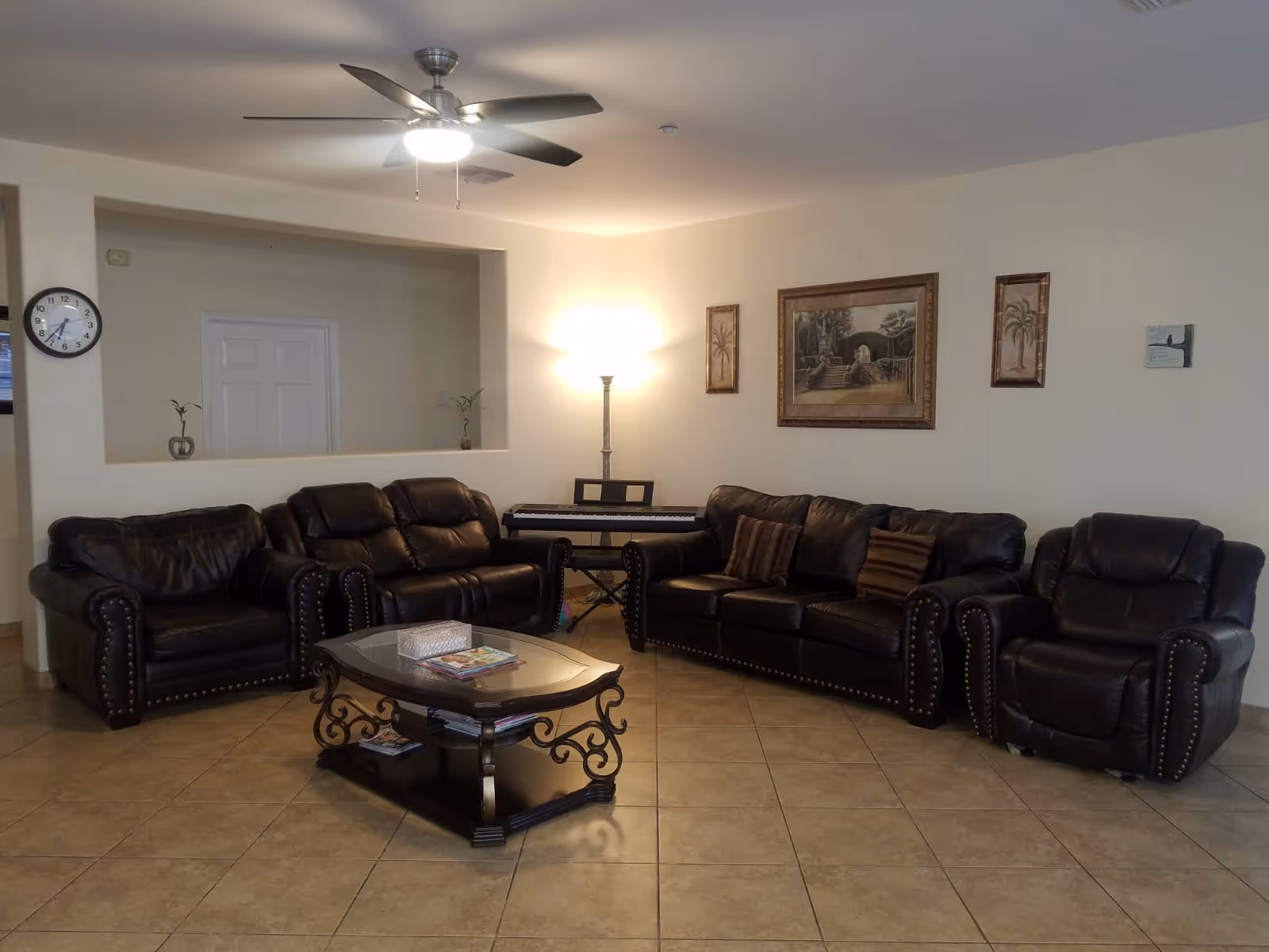 A living room with a set of dark brown leather sofas and armchairs arranged around a glass-top coffee table with ornate legs. The room has beige tiled flooring, a ceiling fan with a light, a floor lamp, and framed artwork on the walls. A digital piano is placed against the wall behind the sofas.