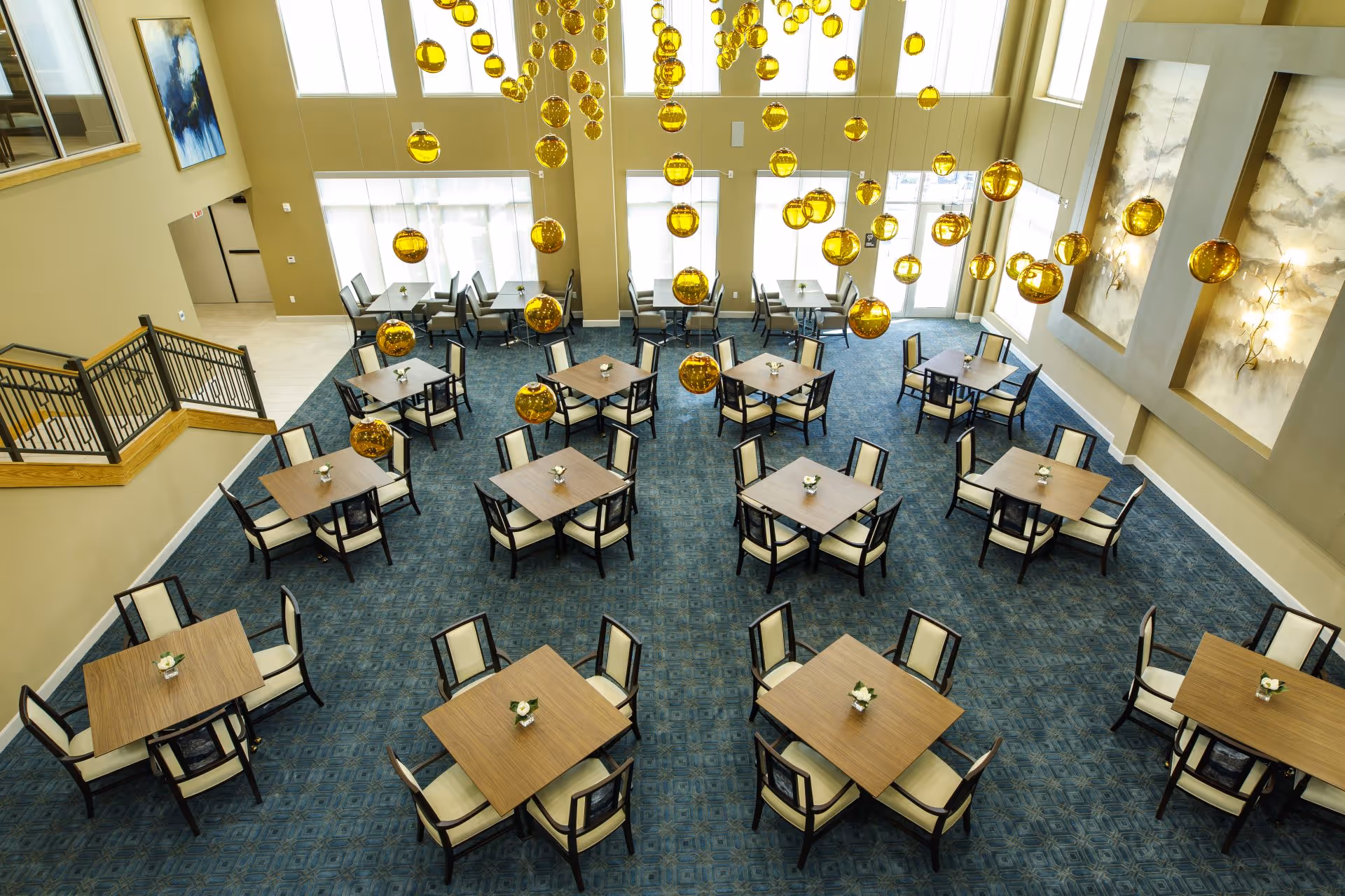 A spacious dining room with multiple square wooden tables, each surrounded by four cushioned chairs. The room features a blue patterned carpet, large windows allowing natural light, and numerous hanging golden spherical light fixtures. There are two large abstract paintings on one wall and a staircase with a wooden and metal railing on the left side.
