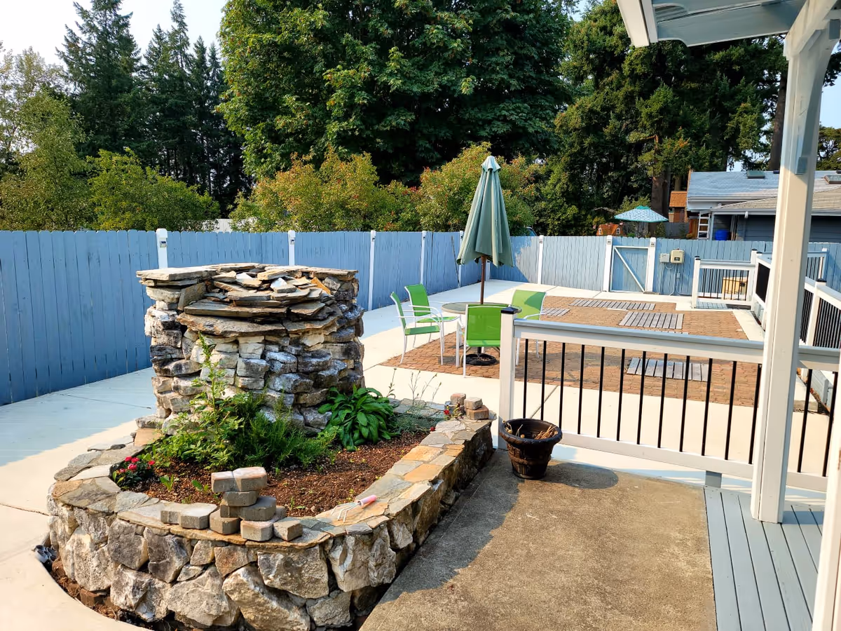 Outdoor patio area with a stone planter and a stone water feature, surrounded by a light blue wooden fence. There is a round table with a closed green umbrella and four green chairs on a paved section. Trees and bushes are visible beyond the fence.