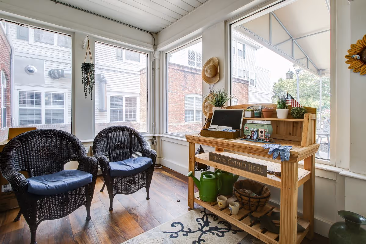 A bright indoor seating area with two black wicker chairs with blue cushions, a wooden table with gardening items including a watering can, gloves, and potted plants. The table has a sign that reads 'Friends Gather Here'. Large windows surround the space, allowing natural light to fill the room and providing views of the building exterior.