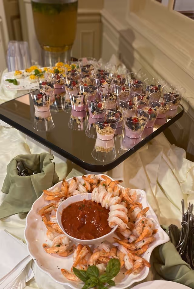 A buffet table with a plate of shrimp arranged around a bowl of cocktail sauce, garnished with basil leaves. Behind it, there is a tray with multiple small dessert cups filled with a pink mousse topped with granola and berries. The table is covered with a cream-colored tablecloth and has green napkins holding utensils.