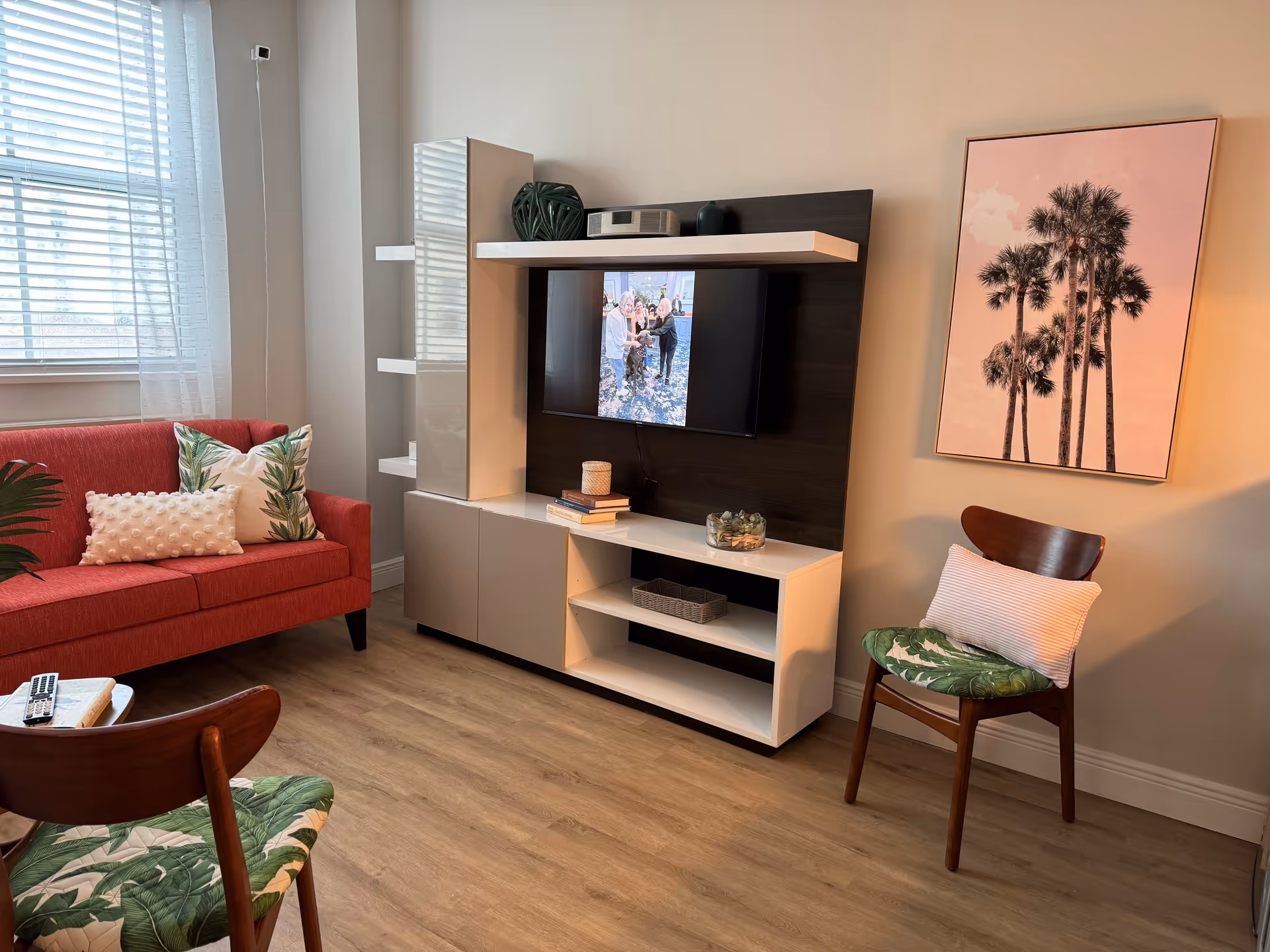 A cozy living room with a red sofa adorned with decorative pillows, two wooden chairs with green leaf-patterned cushions, a modern white and dark wood entertainment unit with a mounted TV displaying a group photo, and a framed picture of palm trees on the wall.