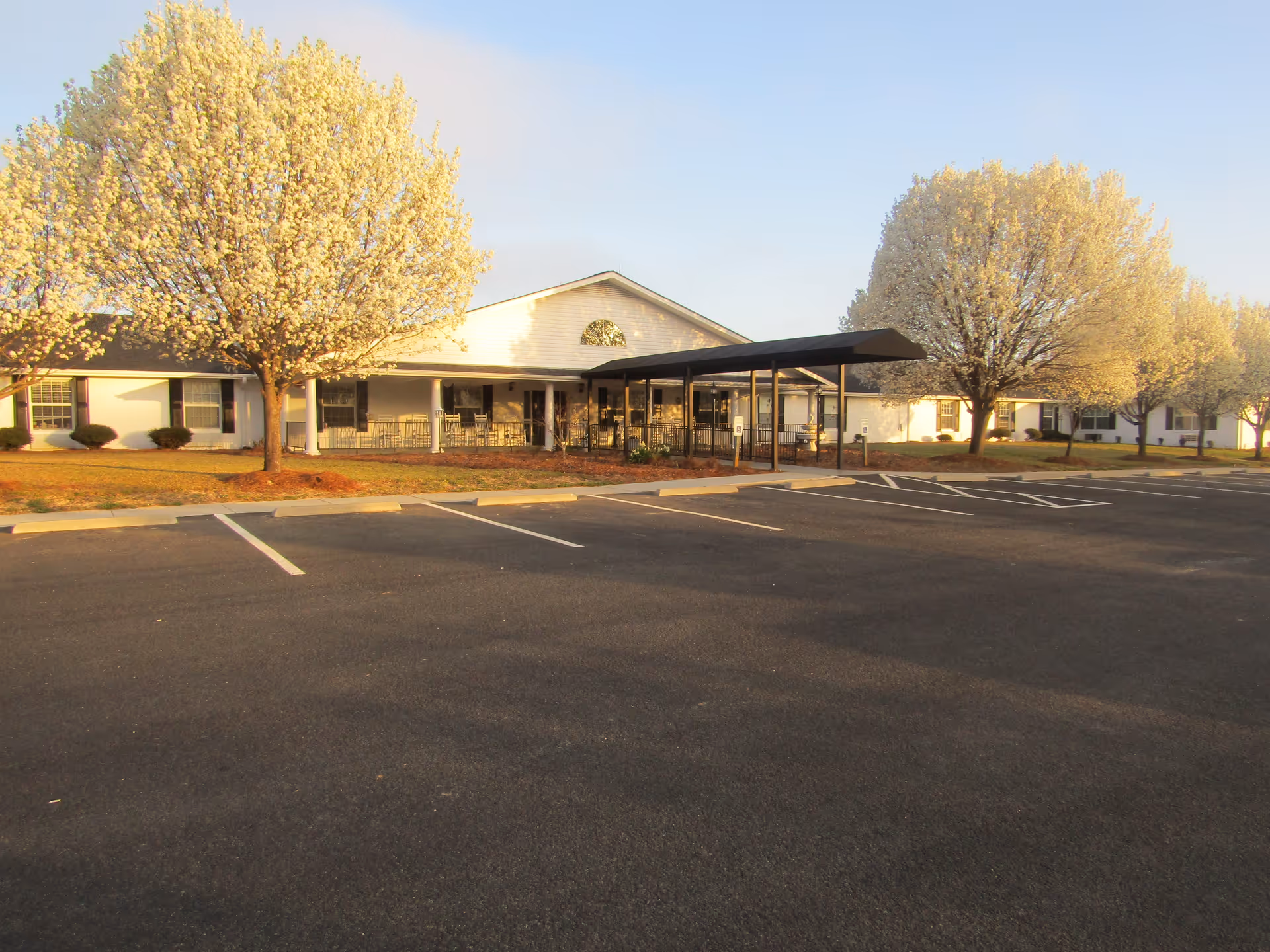 Exterior view of a single-story building with a covered entrance and several blooming trees in front, adjacent to an empty parking lot under a clear sky.