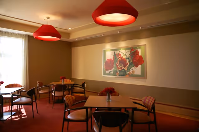 A dining room with several square tables and chairs arranged around them. Each table has a small vase with red flowers. The room has warm lighting with two large red pendant lamps hanging from the ceiling. A large framed painting of red flowers is displayed on the wall. Natural light comes in through a window with sheer curtains on the left side.