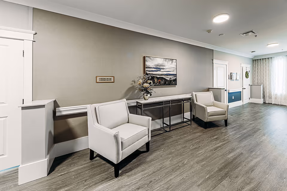 Seating area in a senior living interior hallway with two armchairs, a console table, wall art, and wood-look flooring.