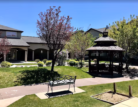 Outdoor garden area at a senior living facility with a black metal bench on a concrete pad, a wooden gazebo, several trees with green and purple leaves, and a building with covered walkways in the background under a clear blue sky.