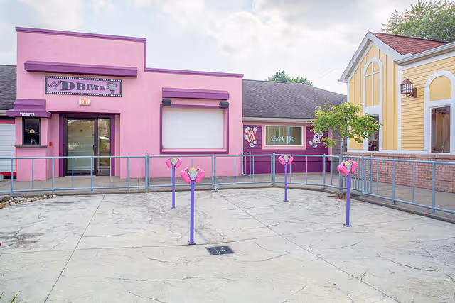 Outdoor view of a colorful drive-in style snack bar area with a pink building labeled 'Drive In' and a purple snack bar window labeled 'Snack Bar'. There are several purple poles with pink holders in front of the buildings, and a yellow building with white trim is visible on the right side. The area is enclosed by a metal railing.