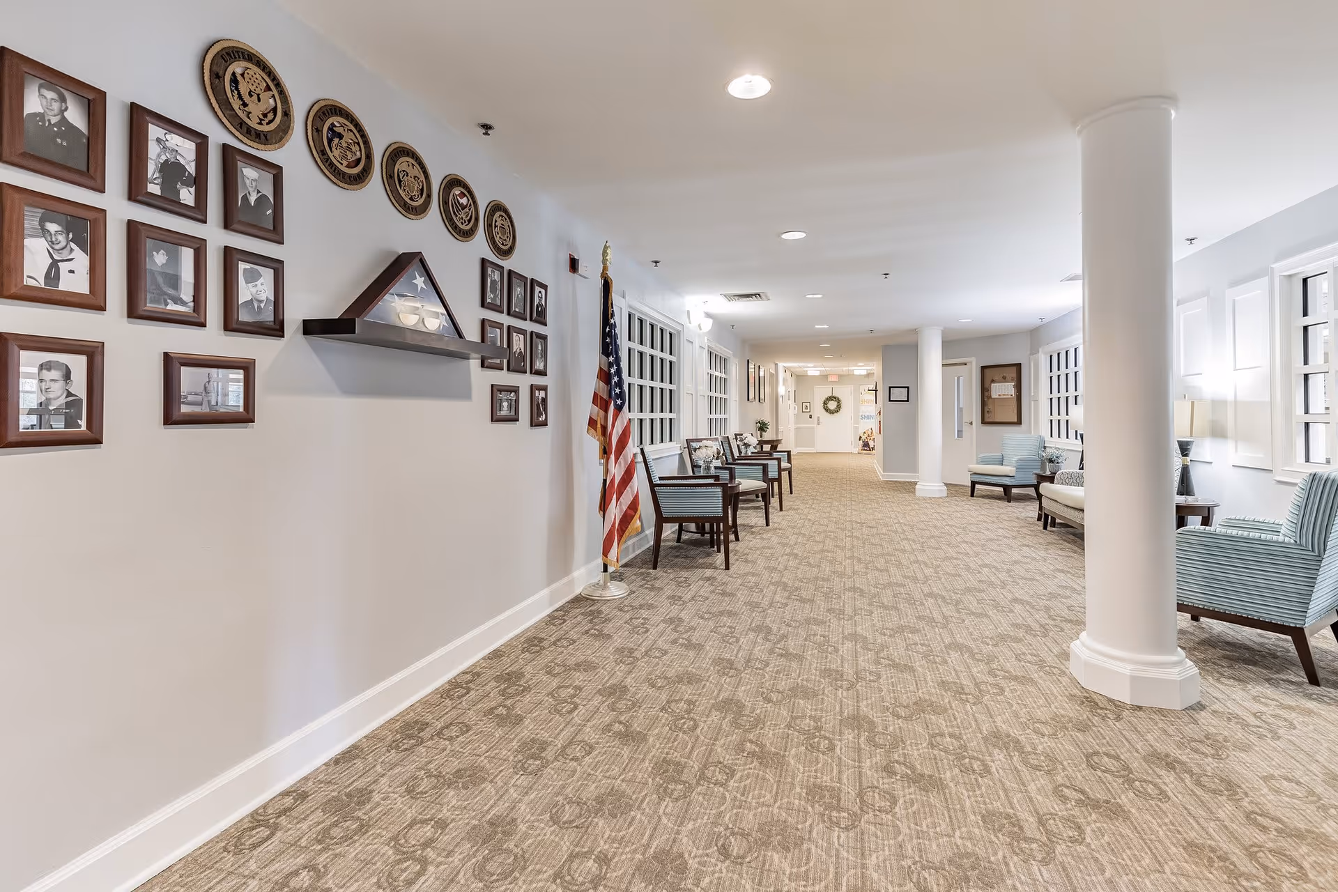 A well-lit hallway in a senior living facility with beige patterned carpet, white walls, and several framed black-and-white photographs and military emblems on the left wall. An American flag stands near a row of wooden chairs with blue cushions along the wall. On the right side, there are more chairs and a small table with a lamp. The hallway extends into the distance with additional seating and decorations visible.