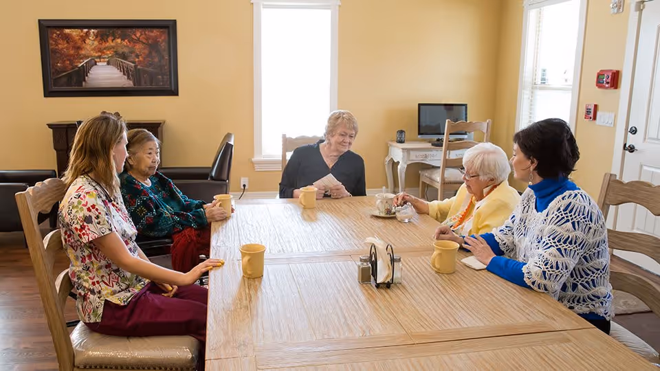 A group of older women and a caregiver sit around a wooden dining table holding mugs in a bright common room.