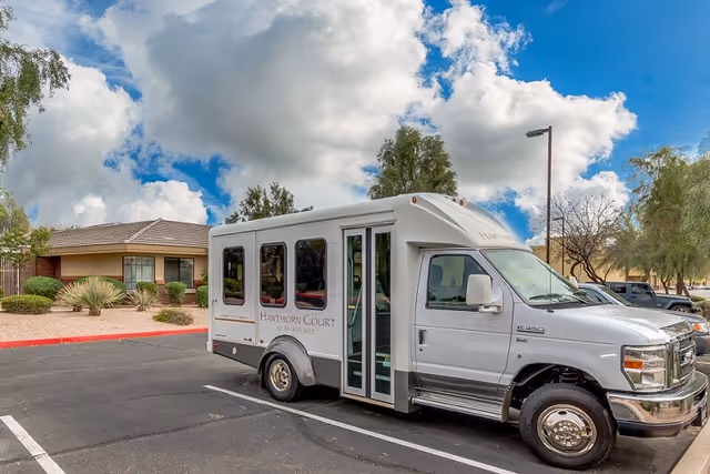 A white shuttle bus parked in a parking lot outside a single-story building with desert landscaping including bushes and small trees under a partly cloudy sky. The bus has the text 'Hawthorn Court at Ahwatukee' on its side.