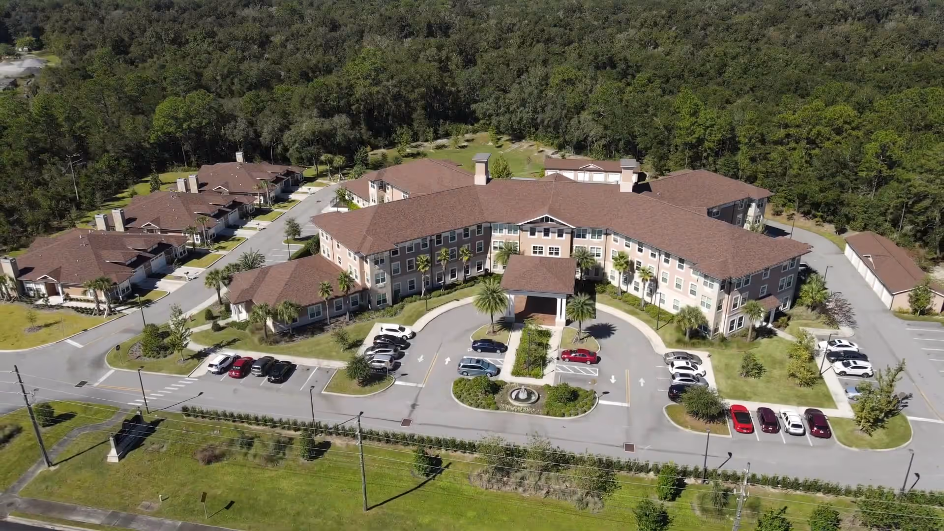 Aerial view of Canterfield of Ocala senior living facility showing a large, multi-wing building with a circular driveway and parking spaces. Surrounding the main building are smaller residential units and a wooded area in the background.