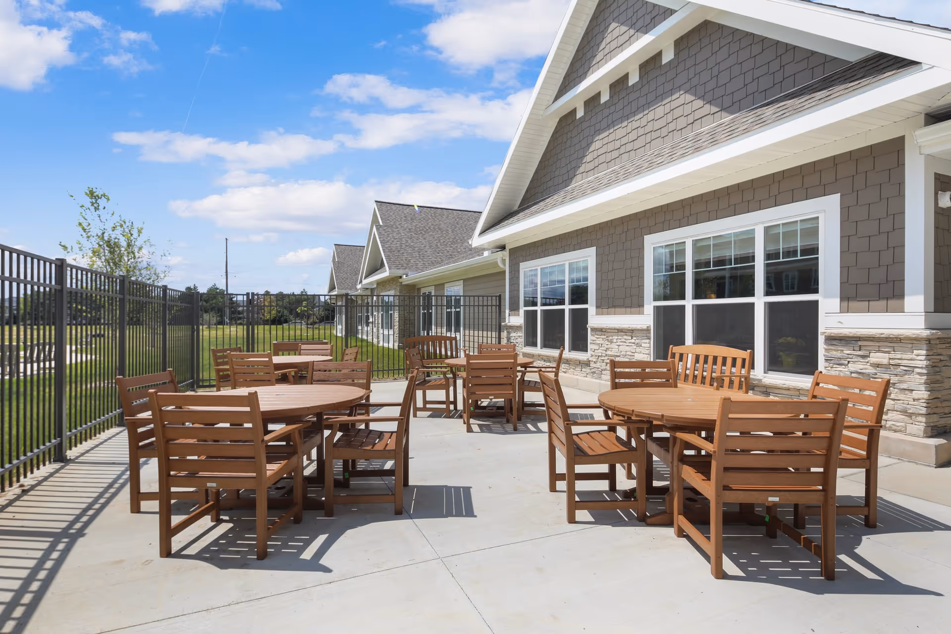 Outdoor patio area with several round wooden tables and matching wooden chairs arranged on a concrete surface next to a building with gray siding and stone accents. The area is enclosed by a black metal fence, and there is a grassy field visible beyond the fence under a partly cloudy blue sky.