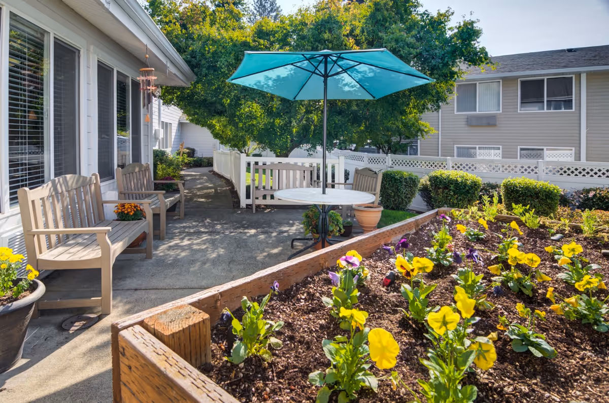Outdoor patio area at a senior living facility with wooden benches, a round table with a blue umbrella, potted plants, and a raised flower bed with yellow and purple flowers. Trees and a white fence are visible in the background.