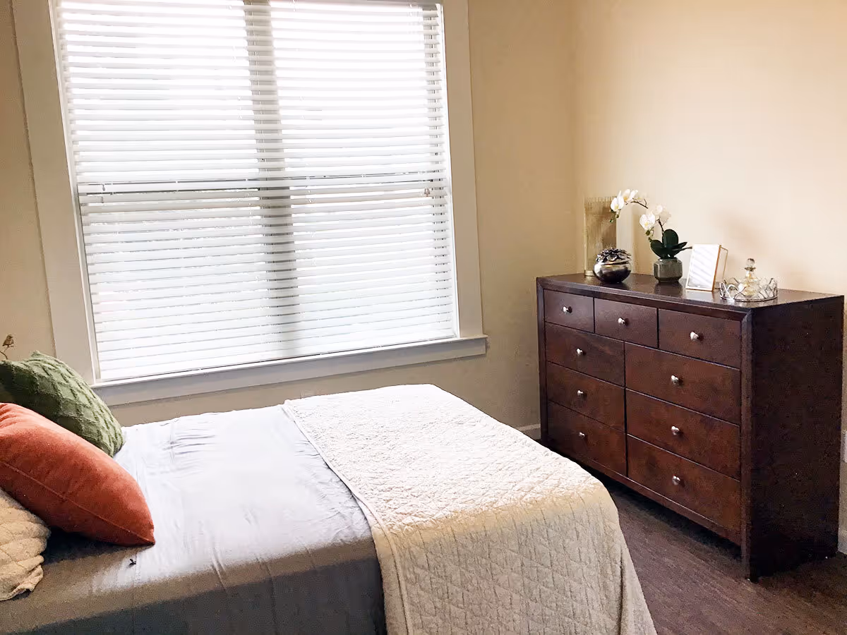 A bedroom with a bed covered in gray and white bedding, accented with green and orange pillows. Next to the bed is a large window with white blinds. Across from the bed is a dark wooden dresser with nine drawers, decorated with a small plant, a framed photo, and other decorative items.