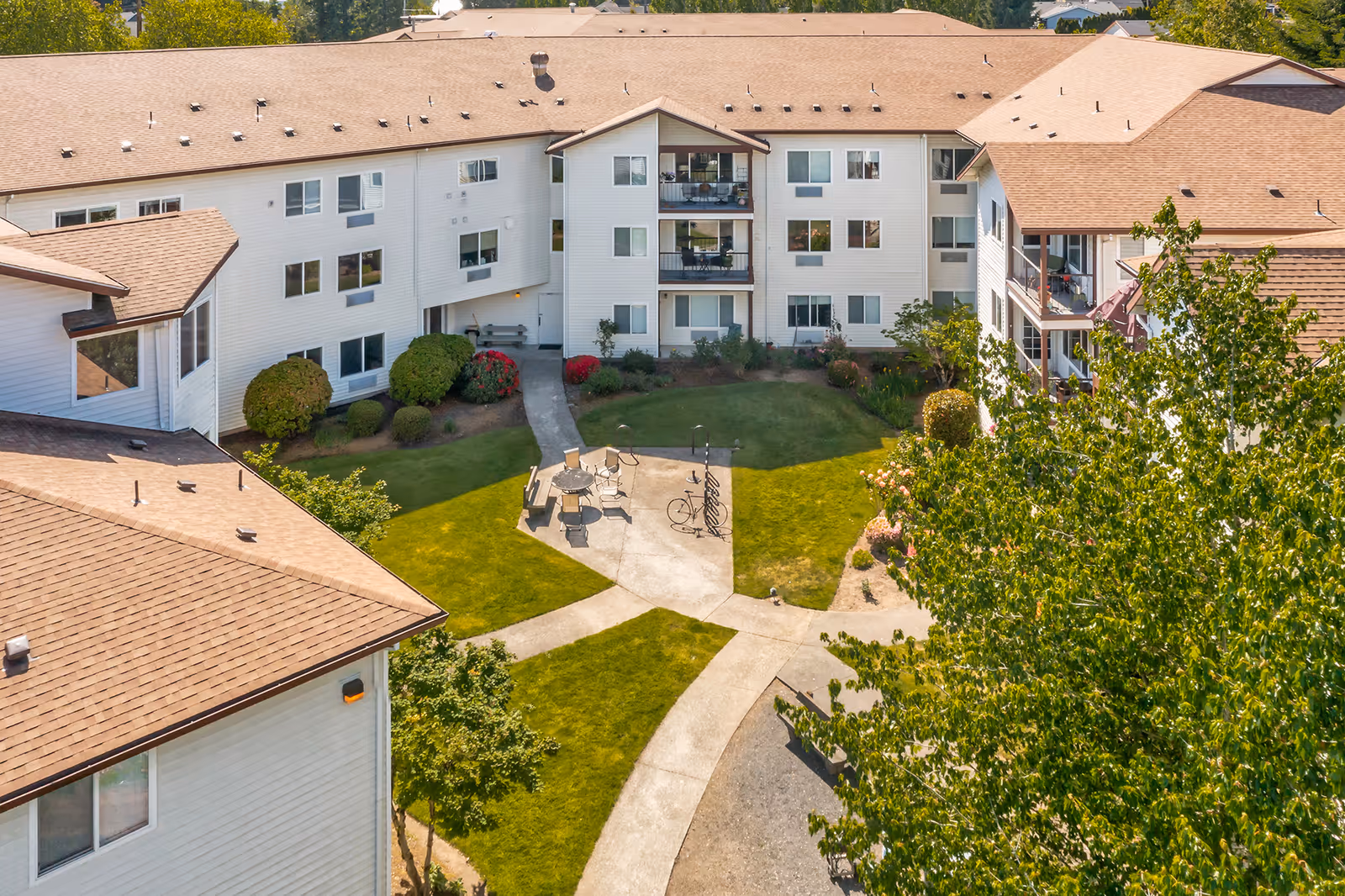 Aerial view of a senior living facility courtyard with a well-maintained lawn, concrete pathways, outdoor seating with tables and chairs, and surrounding white multi-story buildings with balconies and windows. Trees and shrubs are visible around the courtyard.