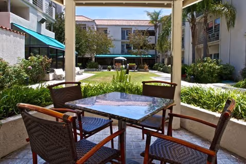 Covered patio table and four chairs overlooking a landscaped central courtyard with lawn, plants, and surrounding multi-story buildings.