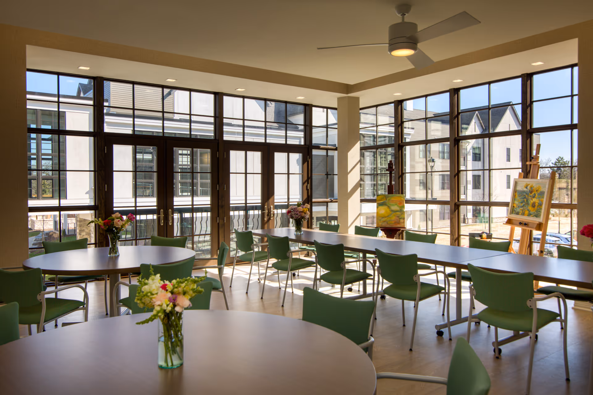 A bright and spacious room with large windows allowing natural light to fill the space. The room contains several round and rectangular tables surrounded by green chairs. Vases with flowers are placed on the tables, and there are paintings on easels near the windows. A ceiling fan is mounted above.