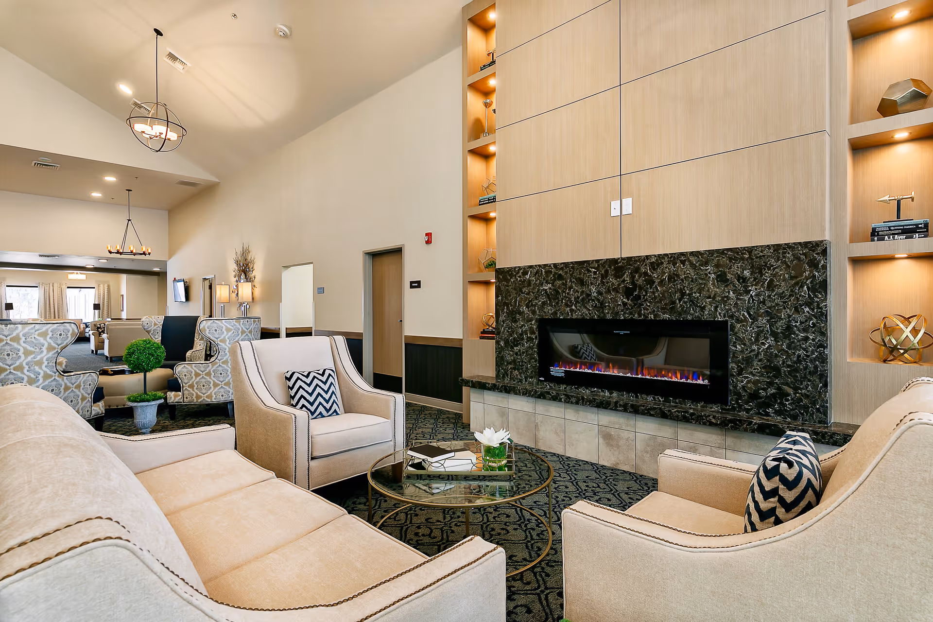 A spacious and well-lit senior living facility lounge area with beige upholstered sofas and armchairs arranged around a glass coffee table. The room features a modern electric fireplace set in a wall with dark marble and light wood paneling, with built-in shelves displaying decorative items. The carpet has a patterned design, and the ceiling has pendant lights. Additional seating and a TV are visible in the background.