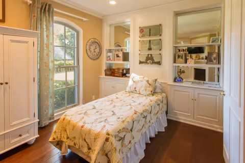 A cozy bedroom with a single bed covered in a floral bedspread. The room features wooden flooring, a large window with floral curtains, built-in white cabinets with shelves displaying decorative items, and a wall clock. The walls are painted in a warm beige tone.